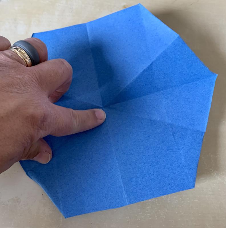 A hand down a piece of blue paper shaped like a regular hexagon, with various creases in it.