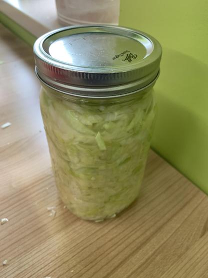 A glass canning jar stuffed with wet green cabbage. On top is a metal lid and rim. The jar sits on a wood table next to a lime green wall. A window sill can be seen in the background.