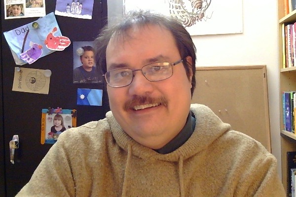 A photo of me, a 44-year-old man, smiling. I'm wearing glasses and a hoodie and have a mustache. Behind me, you can see a small portion of a print of M.C. Escher's "Moebius Strip II", as well as a metal cabinet with a bunch of pictures of my kids, some kids' artwork, and a magnet featuring the Minnesota state flag.